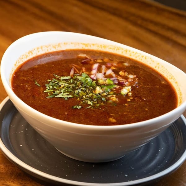 Rich, dark red birria broth in a white bowl, garnished with cilantro, diced red onions, and green chiles.