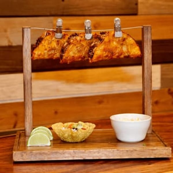 Three tacos hanging from a string on a wooden stand, with lime slices and dips below.