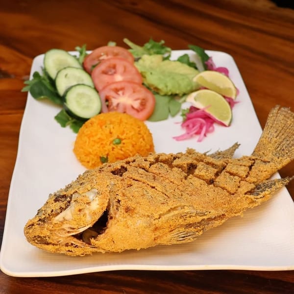 Crispy fried whole fish with rice, sliced tomatoes, cucumbers, avocado, and lime on a white plate.
