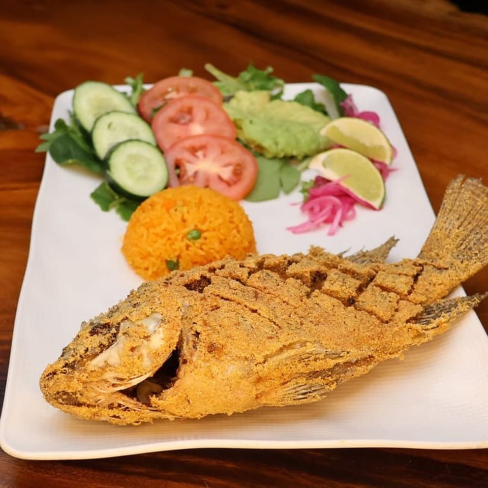 Crispy fried whole fish with rice, sliced tomatoes, cucumbers, avocado, and lime on a white plate.