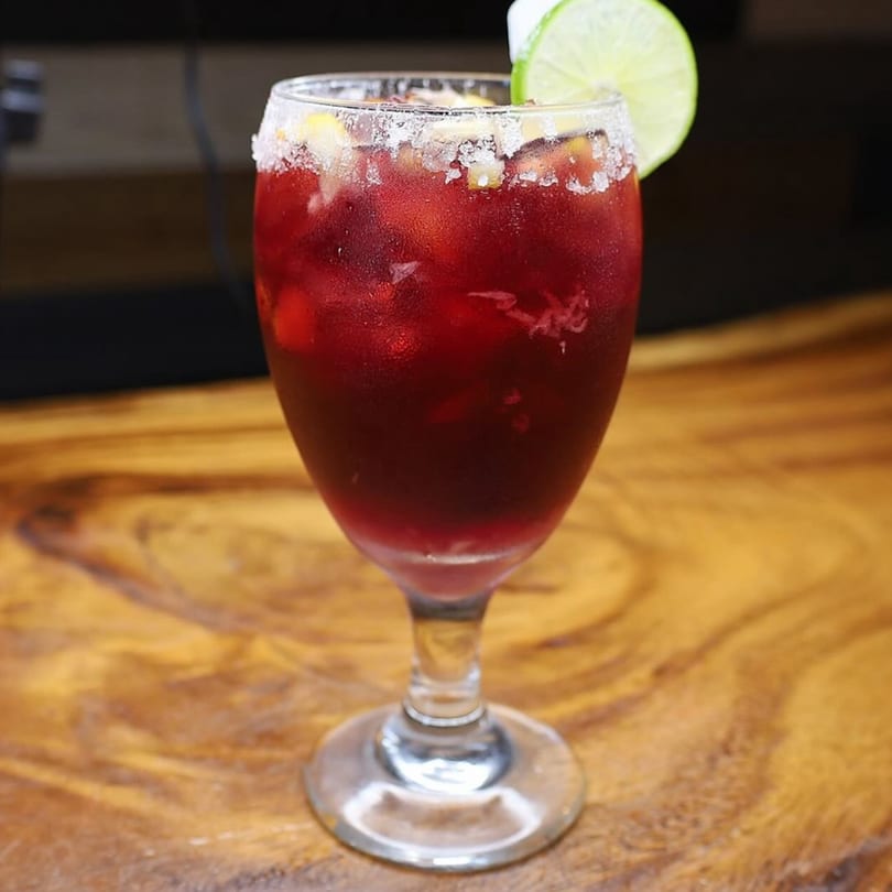 A vibrant red Mexican drink in a stemmed glass, garnished with a lime slice and a salted rim.
