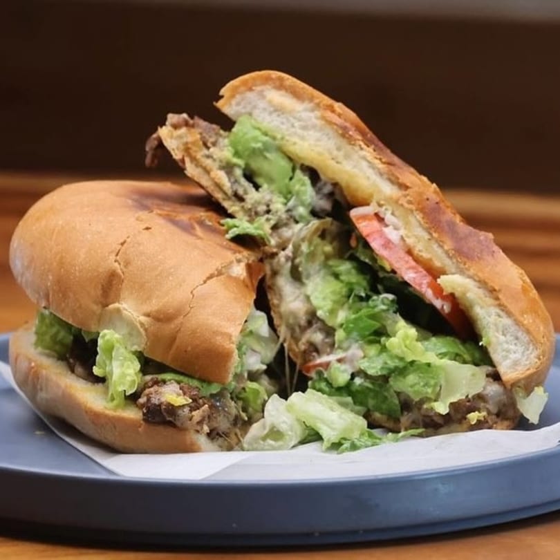 A Mexican torta, cut in half, filled with meat, lettuce, avocado, and tomato on a blue plate.