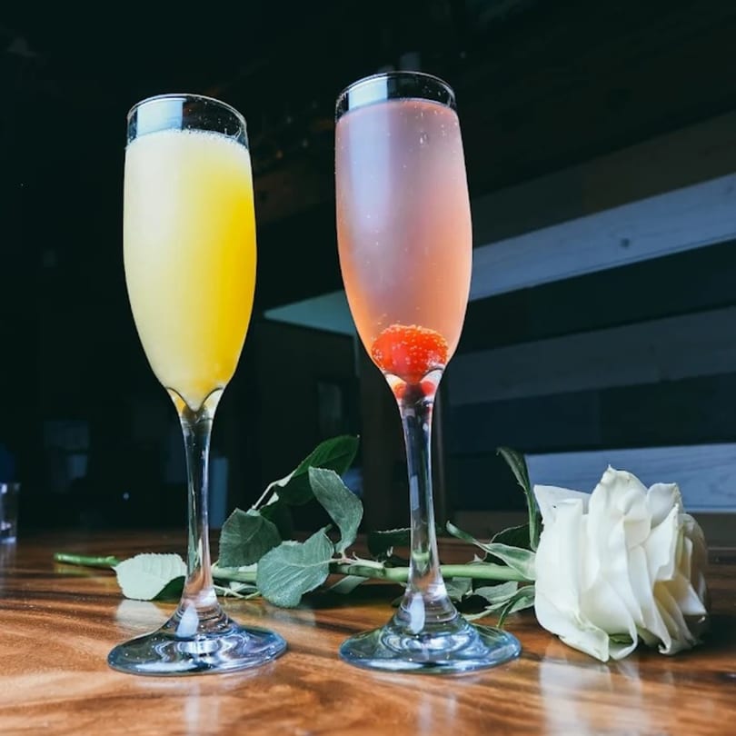 Two champagne flutes, one yellow and one pink with a raspberry, next to a white rose.