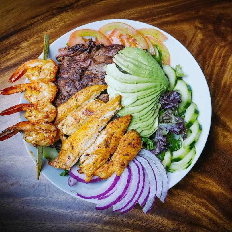 A Mexican dish with grilled shrimp, chicken, steak, avocado, tomatoes, cucumbers, and red onion.
